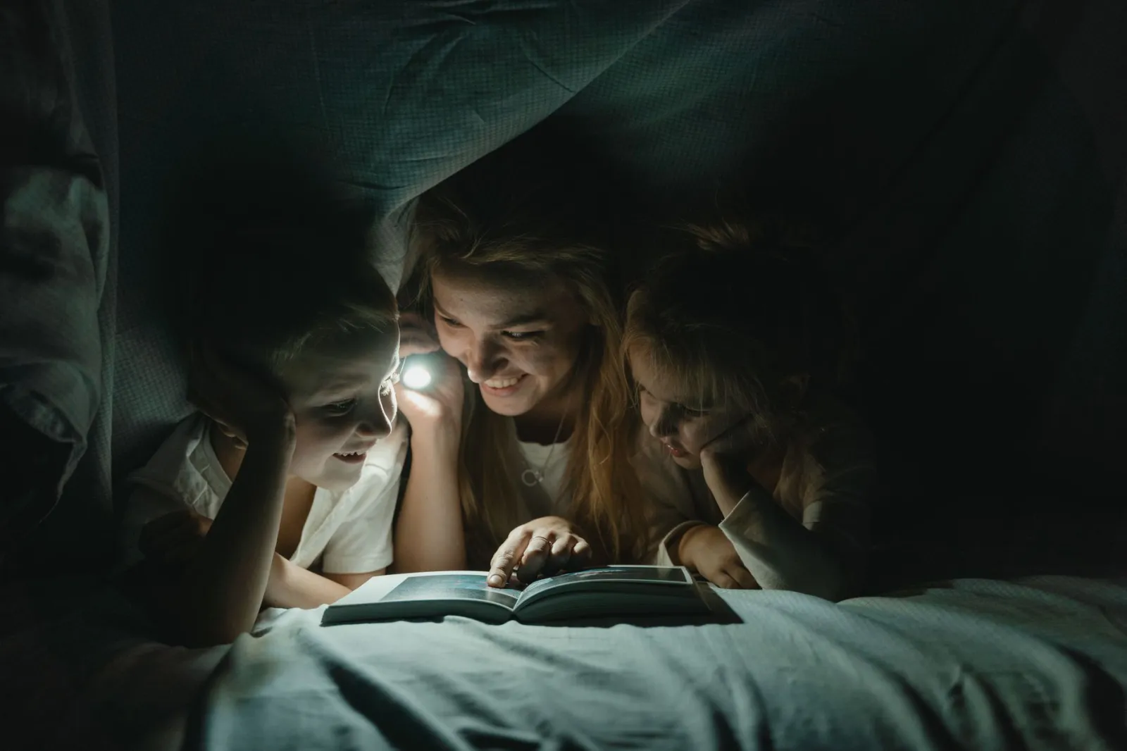A mother reads a bedtime story to her children under the blanket with a flashlight, creating a warm, bonding moment.